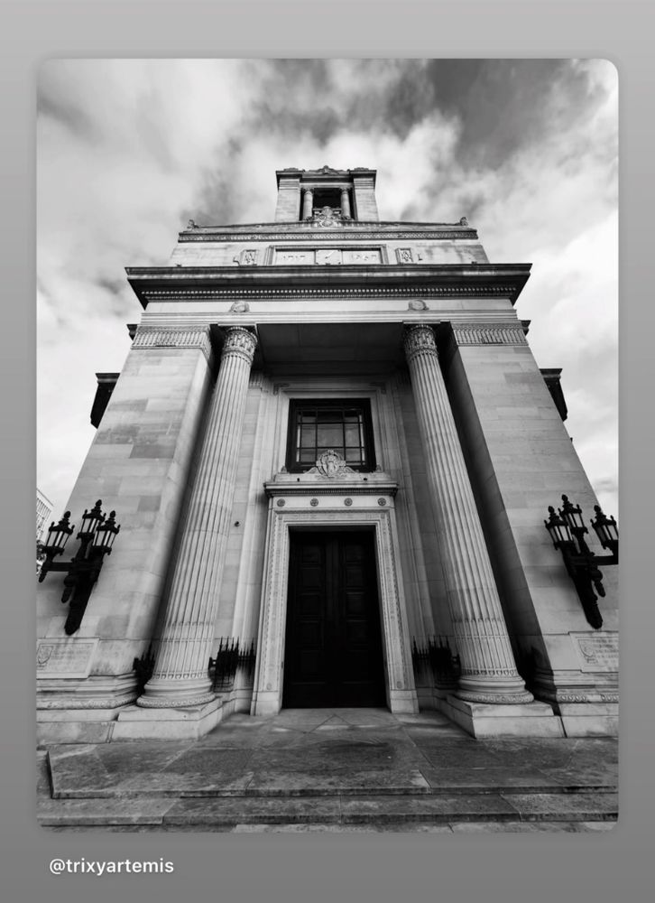 Black-and-white low-angle view of a grand stone building with neoclassical architecture, featuring tall fluted columns, ornate carvings, and large double doors at the entrance. The facade includes decorative lanterns on either side, detailed friezes above the doorway, and a tower-like structure rising at the top. Dramatic clouds fill the sky, enhancing the imposing and monumental appearance.