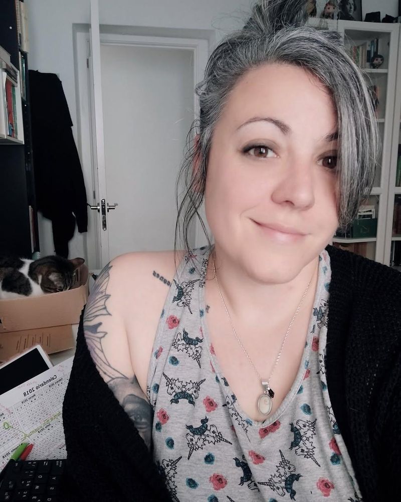 Me smiling, with my hair in a bun and a sleeping cat behind me. On my desk there's a marked 2018 calendar.