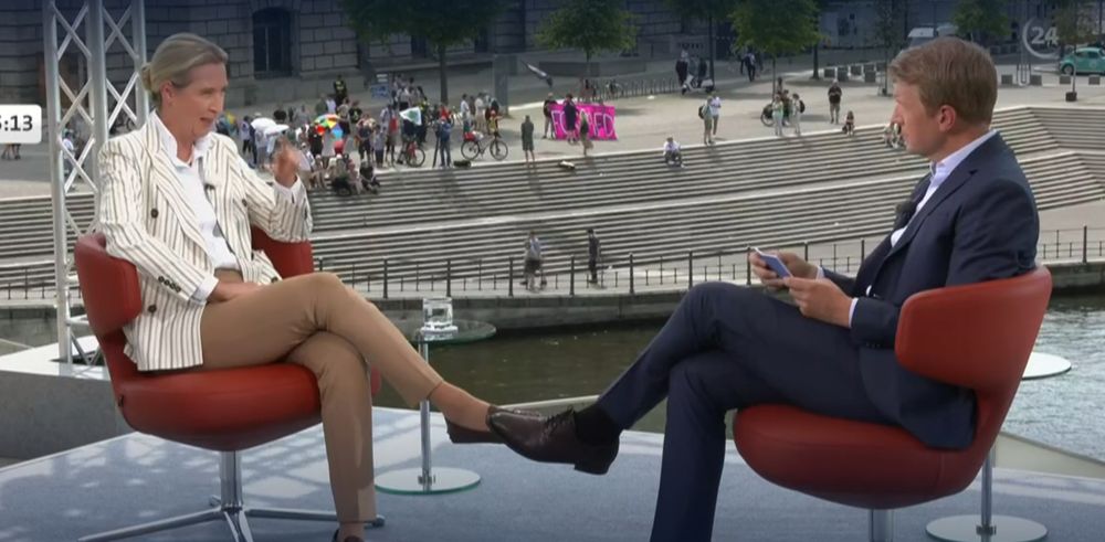 ARD-Sommerinterview. Alice Weidel und Markus Preis; im Hintergrund ist eine kleinere Gruppe Protestierende zu sehen. 