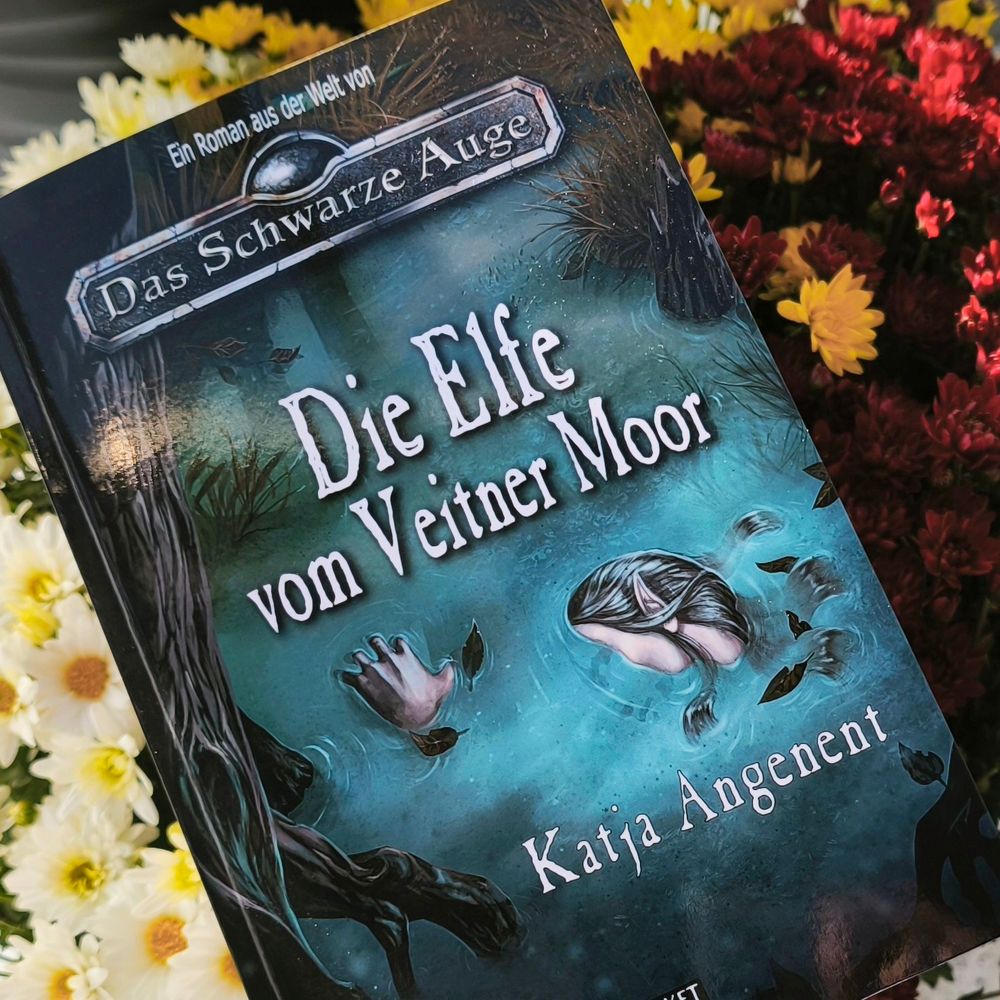 Der Roman "Die Elfe vom Veitner Moor" von Katja Angenent - ein Fantasy-Krimi, der in der fantastischen Welt von Das Schwarze Auge spielt - liegt auf einem Strauß Blumen. 