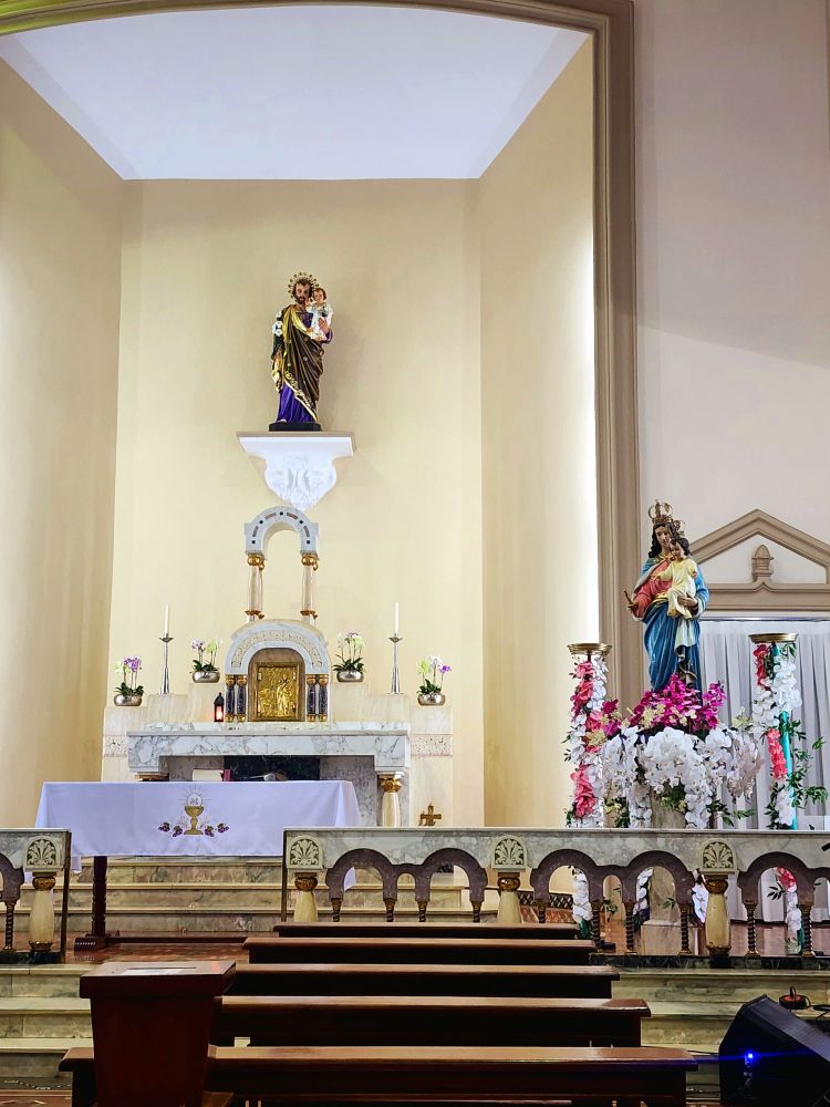 Imagem de um altar em uma igreja católica. Ao centro, na parte de cima uma estátua grande de São José e à direita, uma estátua grande de Nossa Senhora Auxiliadora, enfeitada com orquídeas.