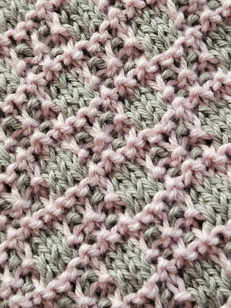 Ein zweifarbiges Strickmuster aus rechten und linken Maschen in einem  Hebemaschenmuster. Hellgraue Quadrate, hellrosa umrandet, wechseln sich ab mit hellgrauen Quadraten, die neben der hellrosa Umrandung auch noch ein hellrosa Kreuz in der Mitte haben.