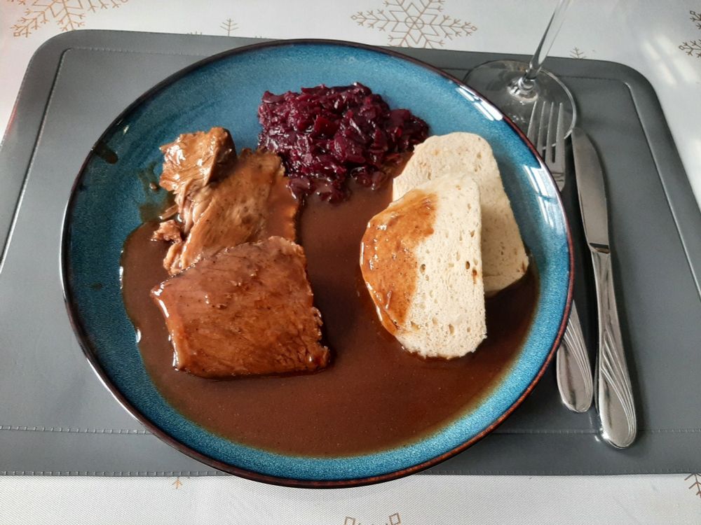 Braten, Schwein und Rind, mit dunkler Soße, Semmelknödel und Rotkraut. Alles auf einen dunkelblauen Teller serviert.