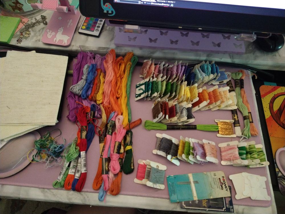 photo of a pile of embroidery floss, some of it in skeins, some on floss bobbins and there's some aida cloth and embroidery needles.