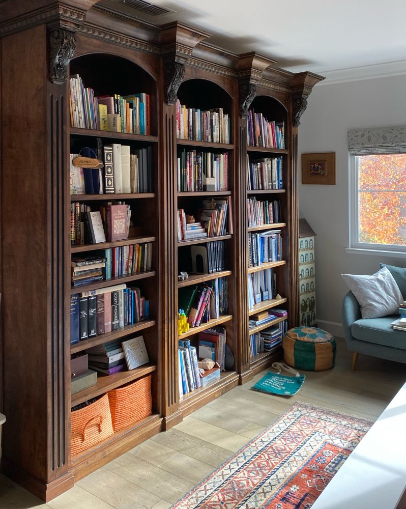 Floor to ceiling bookshelves, wood floor with colorful rug, loveseat in front of window with fall colors outside