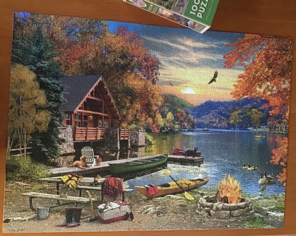 Completed Jigsaw puzzle on a table, scene is of a lakeside cabin surrounded by fall colour trees, there is a pit fire, canoes, dock with two dogs and a bunch of chipmunks around a picnic table. Canada geese at edge of lake, blue hills in background and a sunset sky.