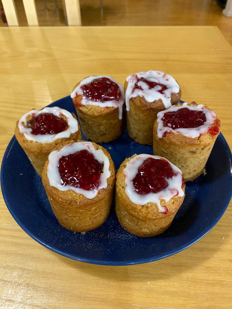 Six gâteaux de Runeberg faits maison sur une assiette, petits cakes à l’amande en forme de cylindres surmontés de confiture à la framboise entourée d’un cercle de glaçage blanc. 