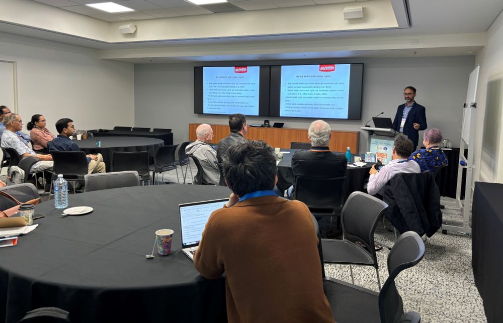Prof Neeraj Gill at a lectern presenting to people sitting at round tables in a workshop