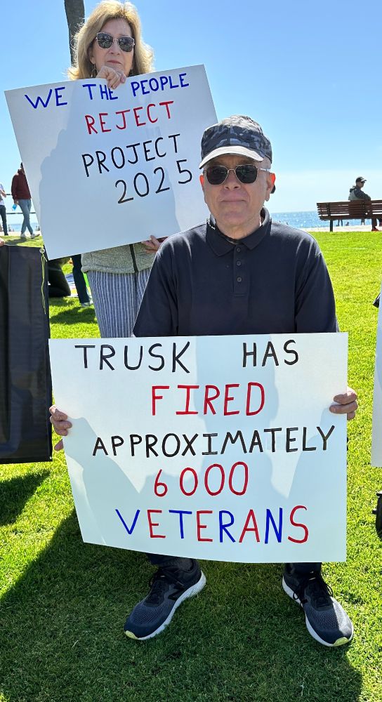 Two people holding protest signs on Main Beach in Laguna Beach CA.  One sign reads: Trusk has fired approximately 6,000 Veterans. 

The second sign reads We the People reject Project 2025