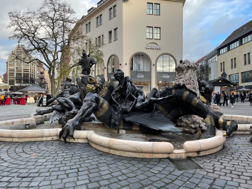 Wider shot of the described sculpture, showing brick pavement and quaint shops around a black metal sculpture