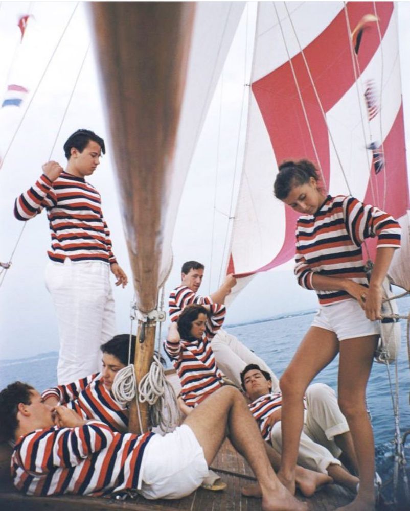 a family of sailors aboard a sailboat with wide striped red/white sails. they all wear short white shorts or white trousers with vibrant red/navy/white striped tops. Several of them are lounging about.