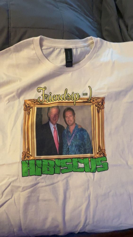 white t shirt flat on bed, print of a golden picture frame, inside is a photo of Bill Clinton next to investor Tom C., on top is text that reads “Friendsip = )” and underneath the frame is a mock logo of the YouTuber Tobuscus, that instead reads “HIBISCUS” in the logo’s font
