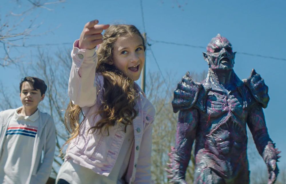 Little girl points towards the camera with her older brother and a large muscular purple alien warlord walking behind her