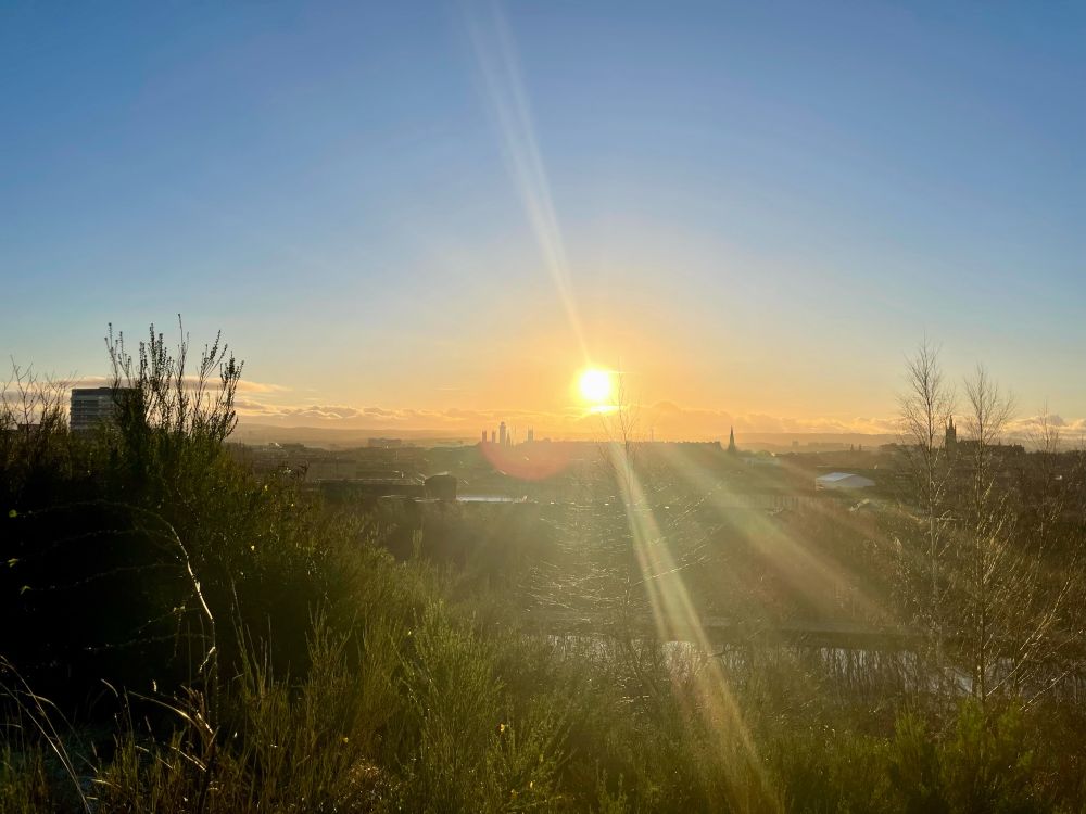 The sun is shining low and bright in a bright blue sky. The Glasgow skyline is visible in the distance
