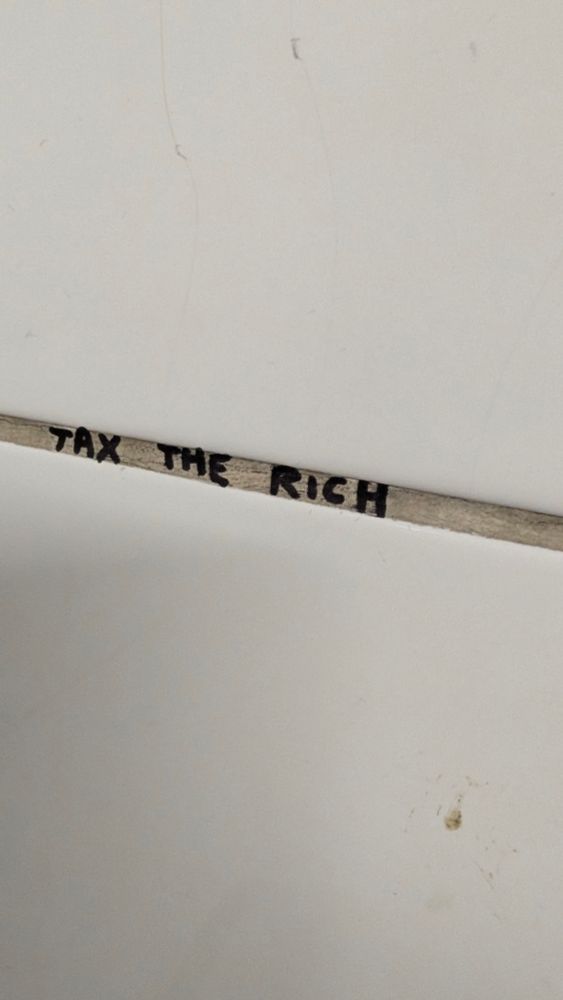 The grout line between white tiles in a slightly grubby pub toilet. Written in the grout line is the statement, "TAX THE RICH"