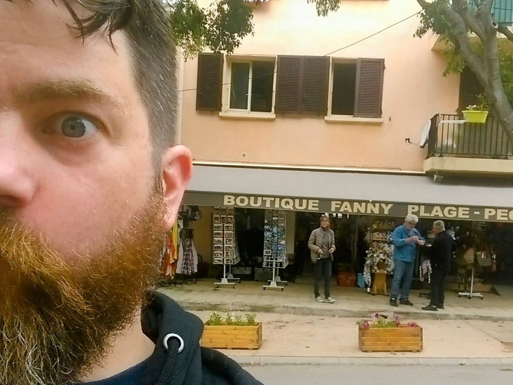 Half of my face covers the left quarter of the photo. My beard looks a little auburn, my hair is brown and trimmed short. A shop behind me on the far side of the street has the name, "Boutique Fanny". I found this funny because I'm a child