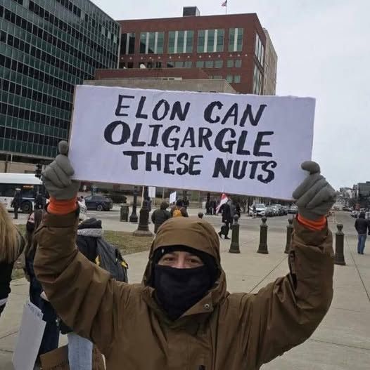 A protester in a brown hood and black face covering holds up a hand written sign which reads, in all capitals, "Elon can oligargle these nuts" 