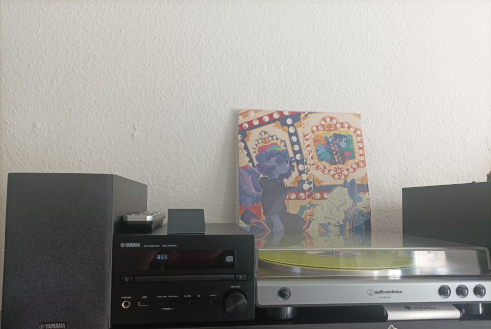 Yamaha receiver with an audio-technica record player plugged in. In it Vylet Pony's Carousel is playing with the cover displayed vertically on top of the record player. Two yamaha speakers complete the black and metallic sound setup that is on top of a black shelf.