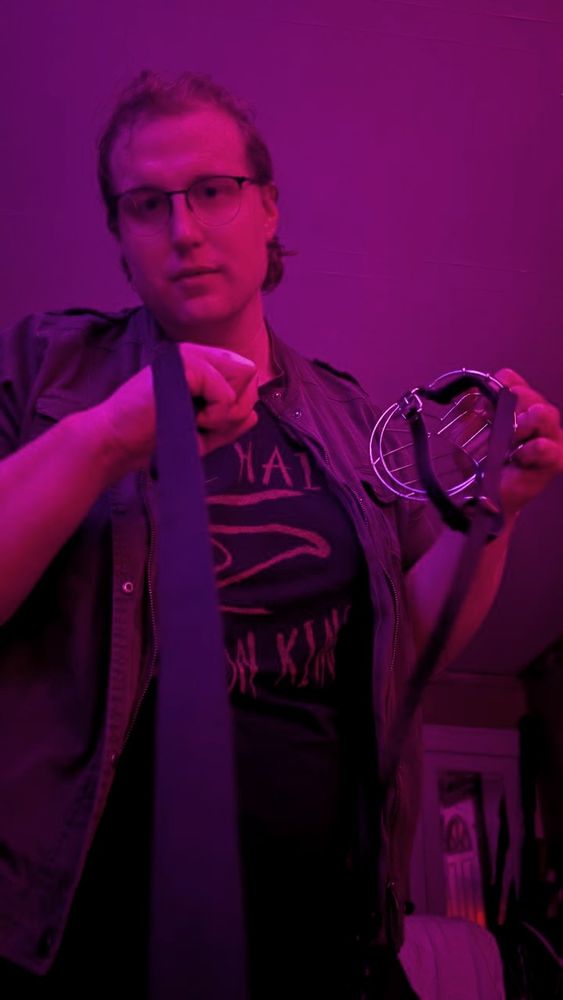 Endless, holding a leash and a muzzle. her hair is pulled back. she is wearing a green jacket and a shirt that reads "All Hail the Crimson King"