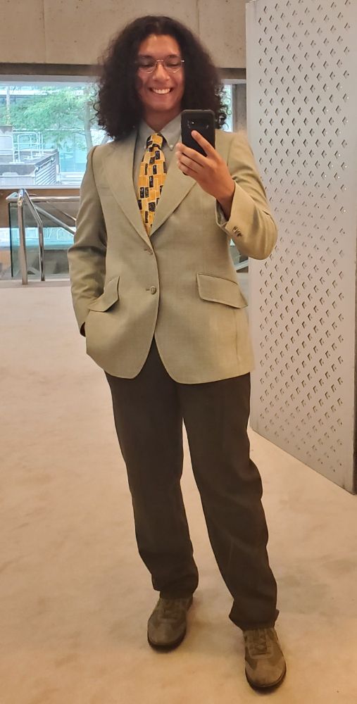 My latino ass in dark green dress pants and boots, paired with a light green suit jacket, and a yellow with blue squares tie. I'm smiling and have my shoulder length black hair down 