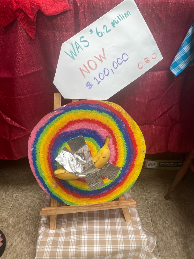 Banana taped to rainbow disc, behind it says “was 6.2 million, now $100,000 or best offer 