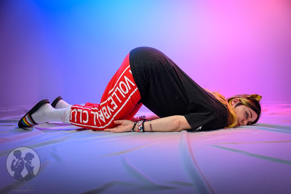 A cosplayer of Kenma from Haikyuu. They have red Nekoma volleyball club sweat pants tucked into white socks and hacve striped slides on. They have a black tshirt and have black to blonde ombre hair, kandi bracelets and Cat ears on. 
