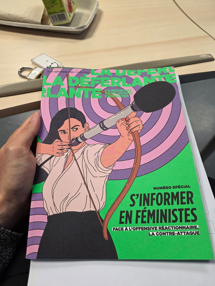 Couverture du numéro 19 de La Déferlante, celui de septembre 2025 sur "S'informer en féministes". L'illustration de couverture est une femme, coupe de cheveux asymétrique long/très courts, qui bande un arc dont la flèche est un microphone géant.
La revue est tenue par une main et en fond, on aperçoit une tablette roulante sur laquelle il y a une petite assiette en carton et une brique de jus de fruit, tout derrière le sol gris d'un hôpital.