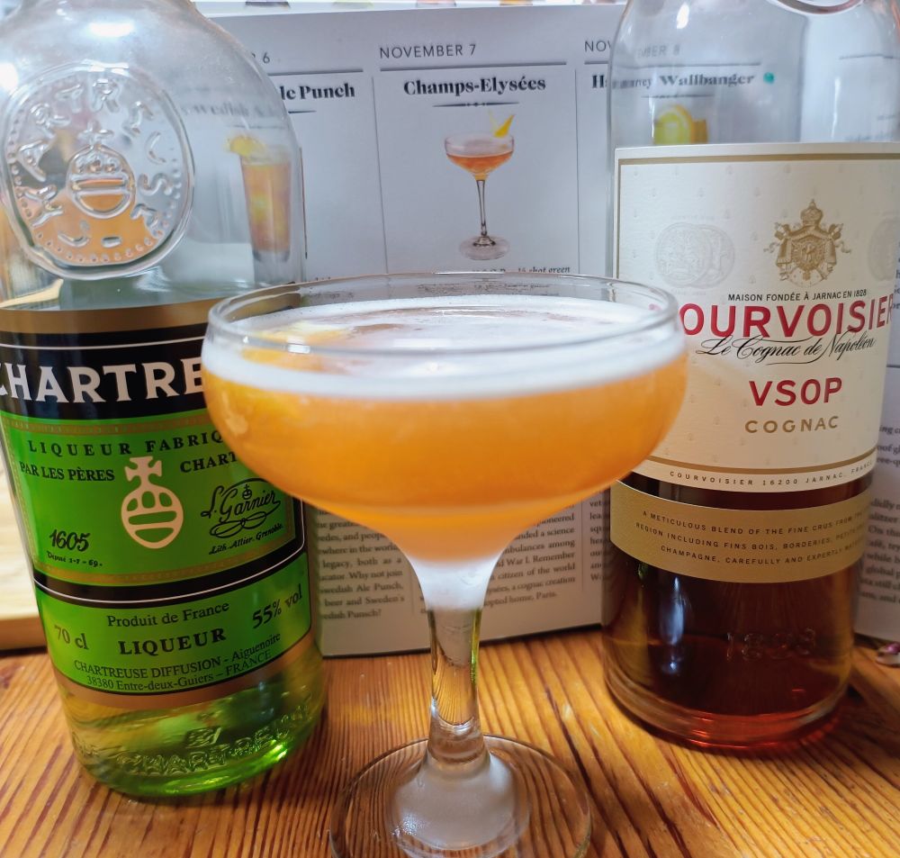 Bottles of green chartreuse and cognac flanking a coupe glass containing a Champs-Elysées cocktail garnished with a lemon twist and all standing in front of the recipe in the Diffords book 365 days of cocktails, which is open and standing up at the back of the image. 