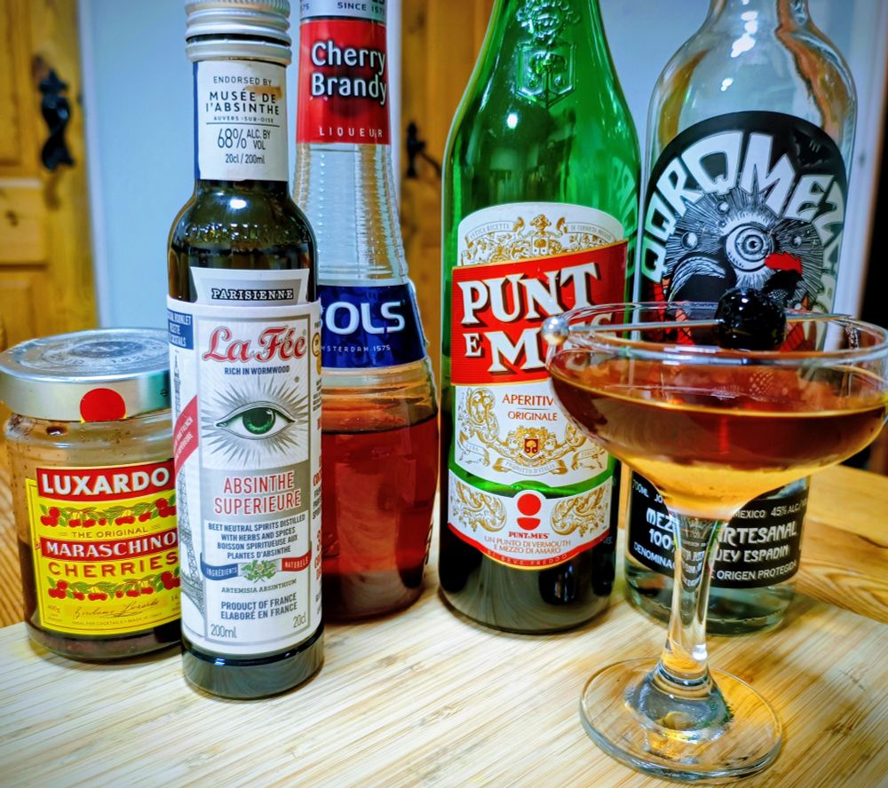 A jar of Maraschino cherries next to a row of bottles containing absinthe (small bottle), cherry brandy, punt e mes and Mezcal (regular sized bottles) and - to the right end of the row - a coupe glass containing an amber coloured cocktail garnished with a Maraschino cherry on a pick. 