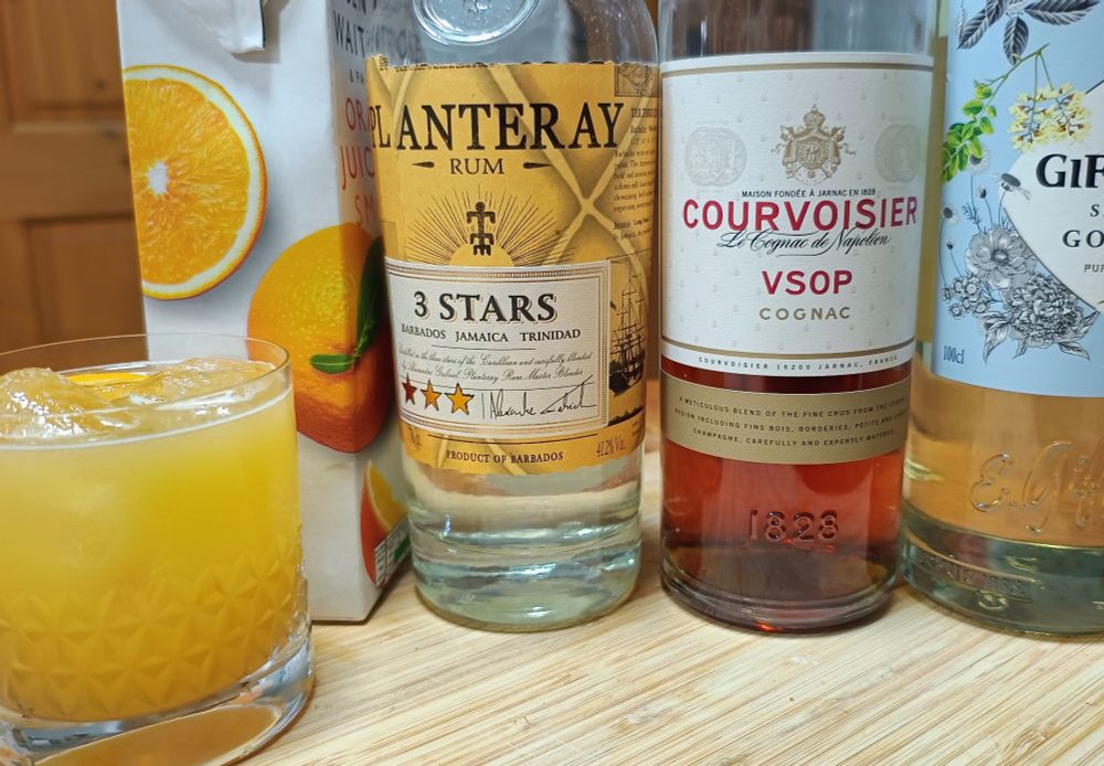 A carton of orange juice next to bottles of light rum, cognac and sugar syrup, all lined up behind a rocks glass containing a Sputnik cocktail, over ice and garnished with an orange slice. 