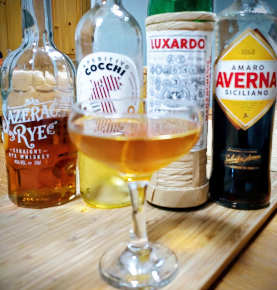 Bottles of rye whiskey, Cocchi Americano, Maraschino Liqueur and Averna amaro in a row behind a coupe glass holding a variation on a Bensonhurst cocktail garnished with an orange twist. 