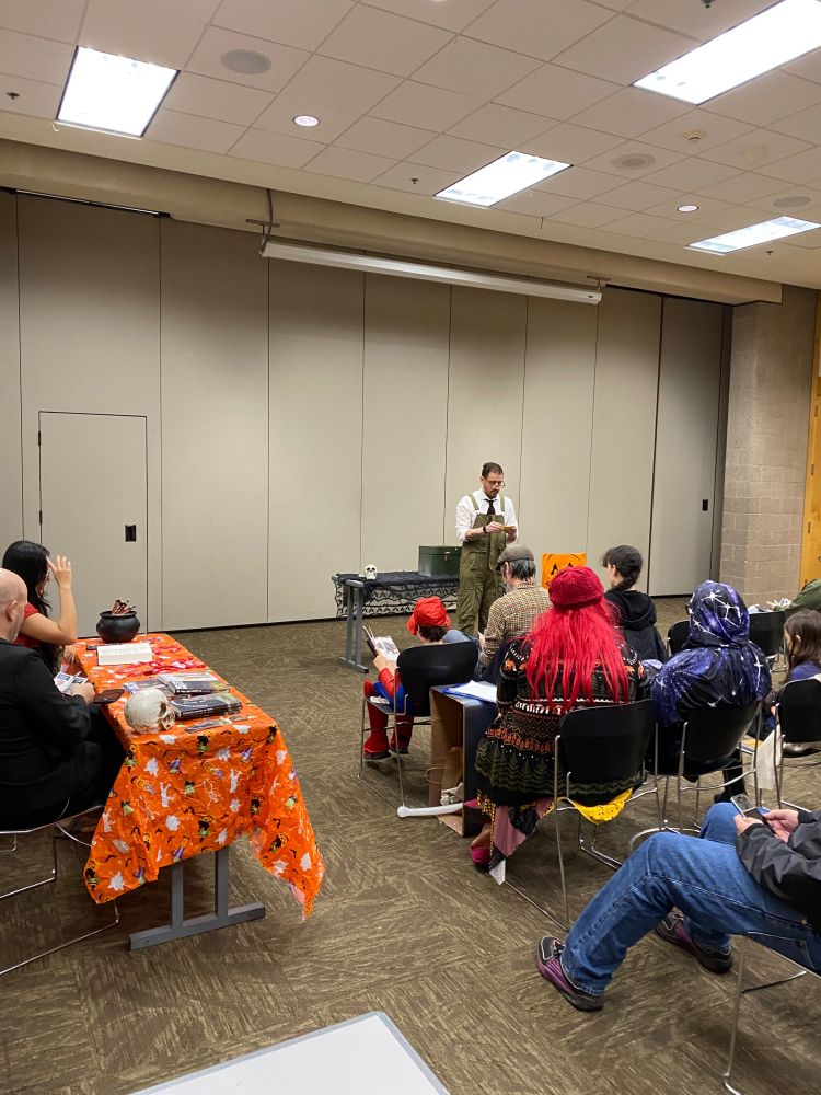 J.B. Kish instructs the audience on proper monster feeding protocols at the Horror Authors Book Party at Beaverton City Library on Saturday, October 25, 2025. The monster sits on a table behind him, in a green metal box.
