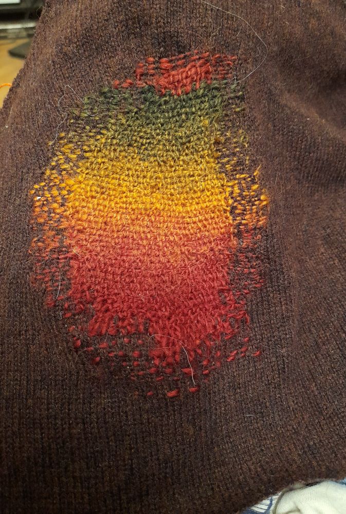Dark brown machine knitted fabric from a jumper. A large hole in it has been mended with rows of blanket stitches in autumn colours which fade into each other.