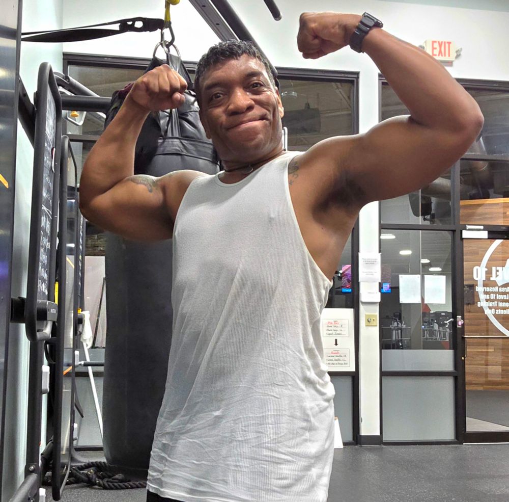 Caleb, wearing a white tank top, flexes in front of a black heavy bag