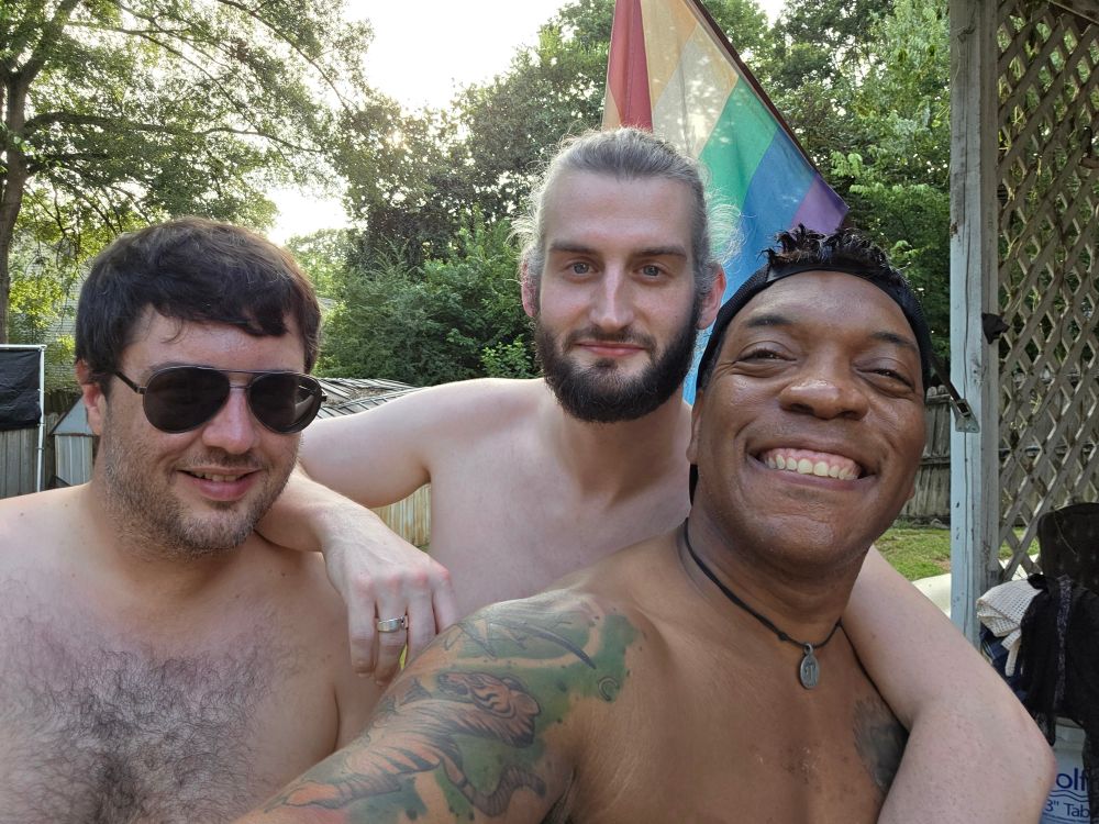 Caleb poses shirtless with his friends in front of a pride flag.