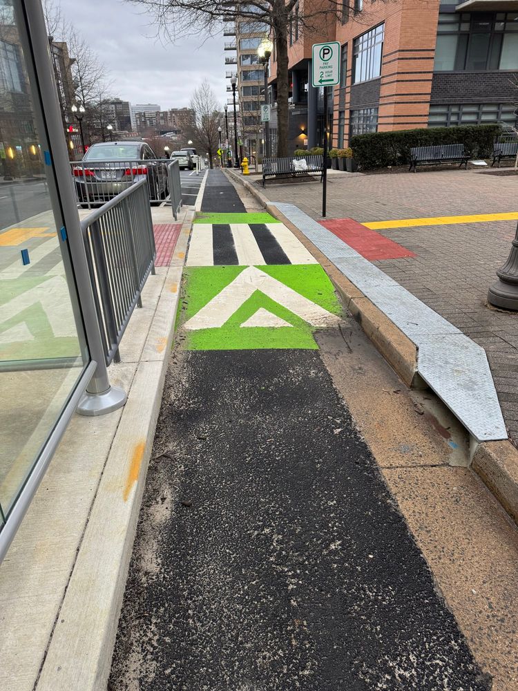 A protected bike lane between the sidewalk and a floating bus stop near 2000 Clarendon Blvd in Arlington, VA. The bike lane ramps up to meet the pedestrian crossing between the sidewalk and bus stop. There is a gutter on on the right side of the bike lane. At the beginning of the ramp, water is diverted from the gutter into a channel directly behind the curb. The channel is covered with metal panels. The channel continues behind the curb to the other side of the pedestrian crossing where water is directed back to the gutter in the bike lane. 