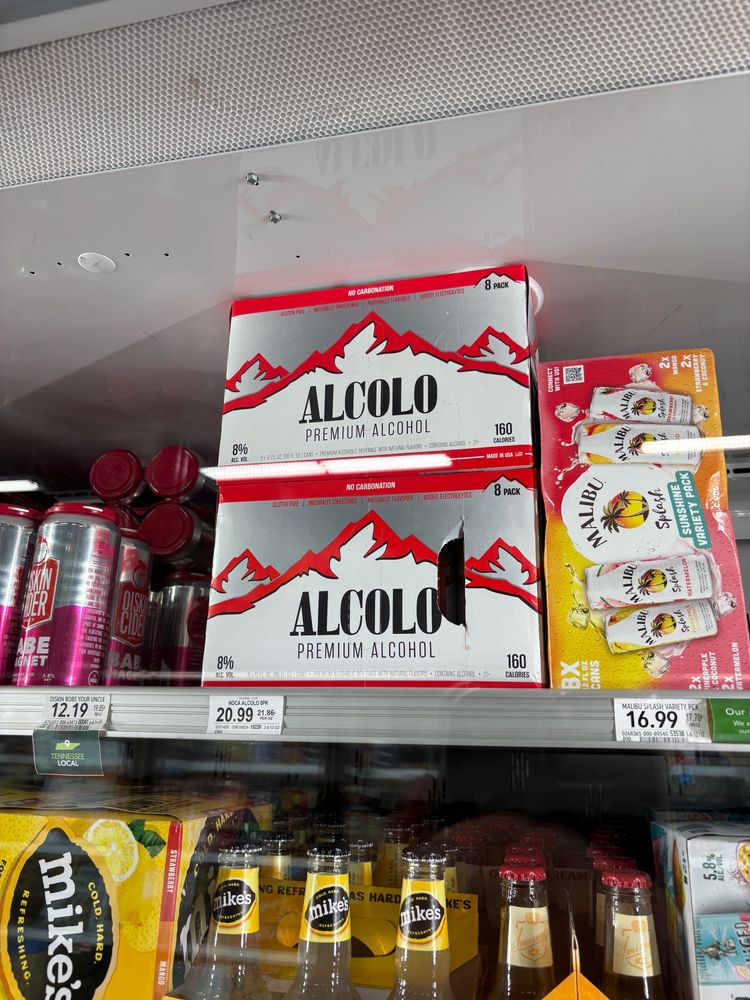 A picture of a beer fridge in a grocery store in America. The subject is a box of beer called Alcolo Premium Alcohol. The box is red and white and silver and the background is some mountains. The text reads: VOL. 8% 
NO CARBONATION
NATURALLY SWEETENED/ NATURALLY FLAVORED ADDED ELECTROLYTES
8 PACK
ALCOLO
PREMIUM ALCOHOL
160 calories
8x 12 FL. O2 (86 FL. OZ.) CANS • PREMIUM ALCOHOLIC BEVERAGE WITH NATURAL FLAVORS • CONTAINS ALCOHOL • 21+
—
