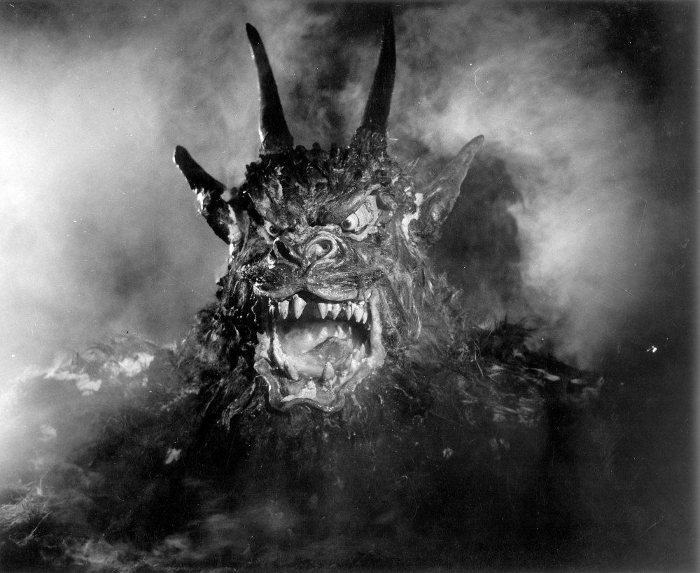 demon from Night of the Demon. close up on a very obviously fake, some would say "goofy" monster with horns and big teeth and fog behind him.