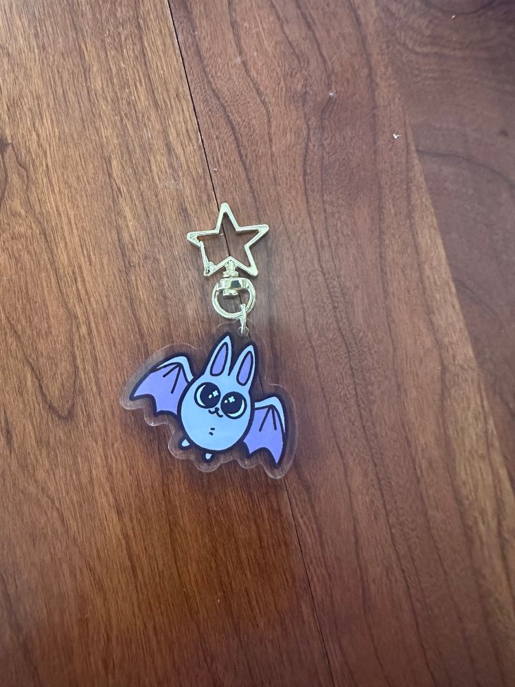 An acrylic keychain of a smiling purple cartoon bat. 
