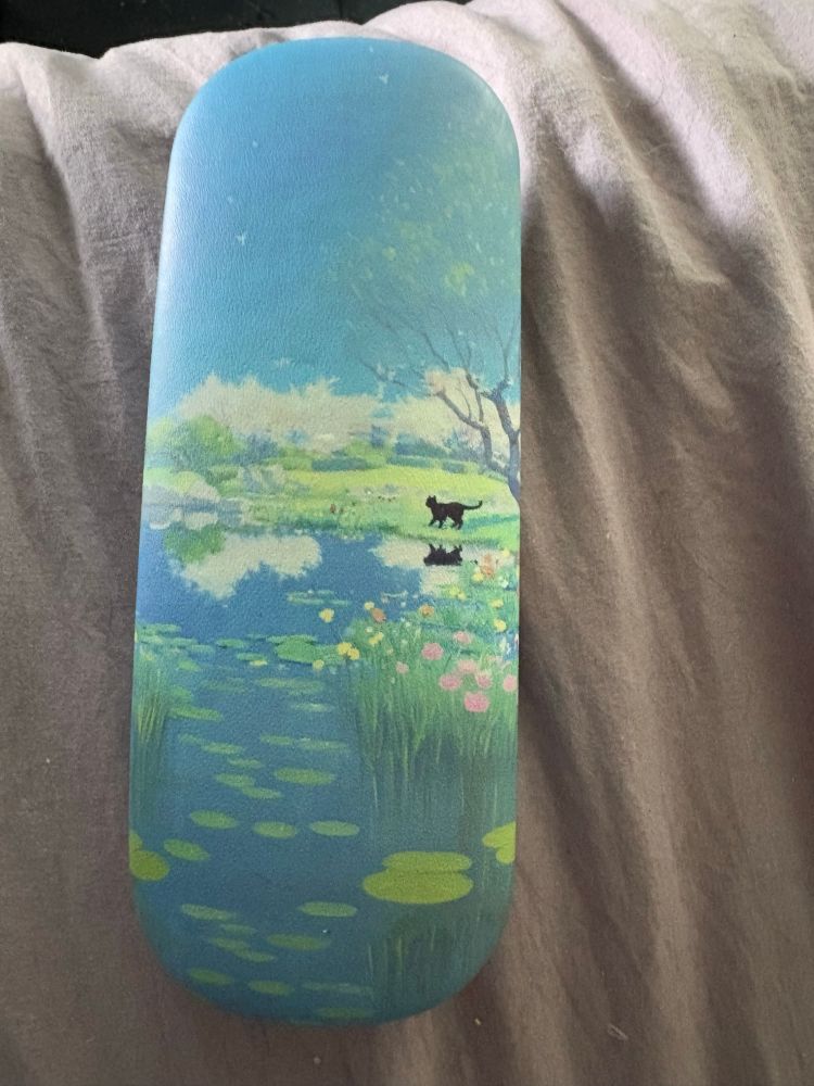 A glasses case that loooks like a painting from Monet or maybe the chili movie Howls Moving Castle. It is a scene of a pond with lily pads, bordered by flowers, and a tree under which a black cat is looking across the water