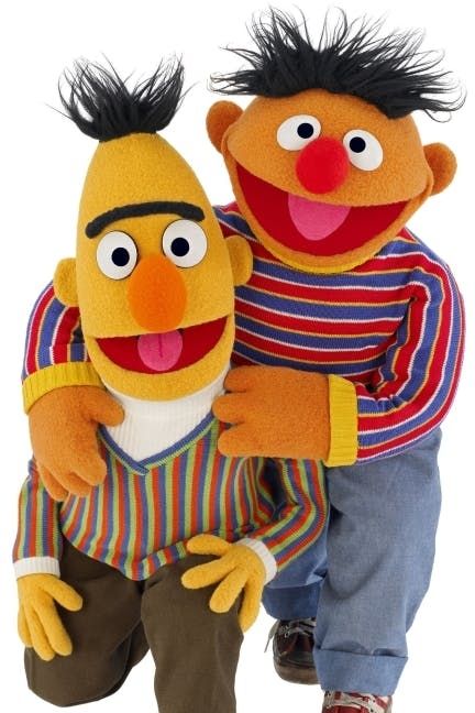 Two Muppets, Bert and Ernie, are facing the camera and smiling. Bert is wearing an orange, blue, and green vertical striped long sleeve shirt over a white turtleneck, and Ernie is wearing a red, blue, and white horizontally striped long sleeved shirt with a yellow collar and cuffs. Bert is kneeling on one knee and Ernie is crouched with his arms around Bert. Both are looking at the camera and smiling. 