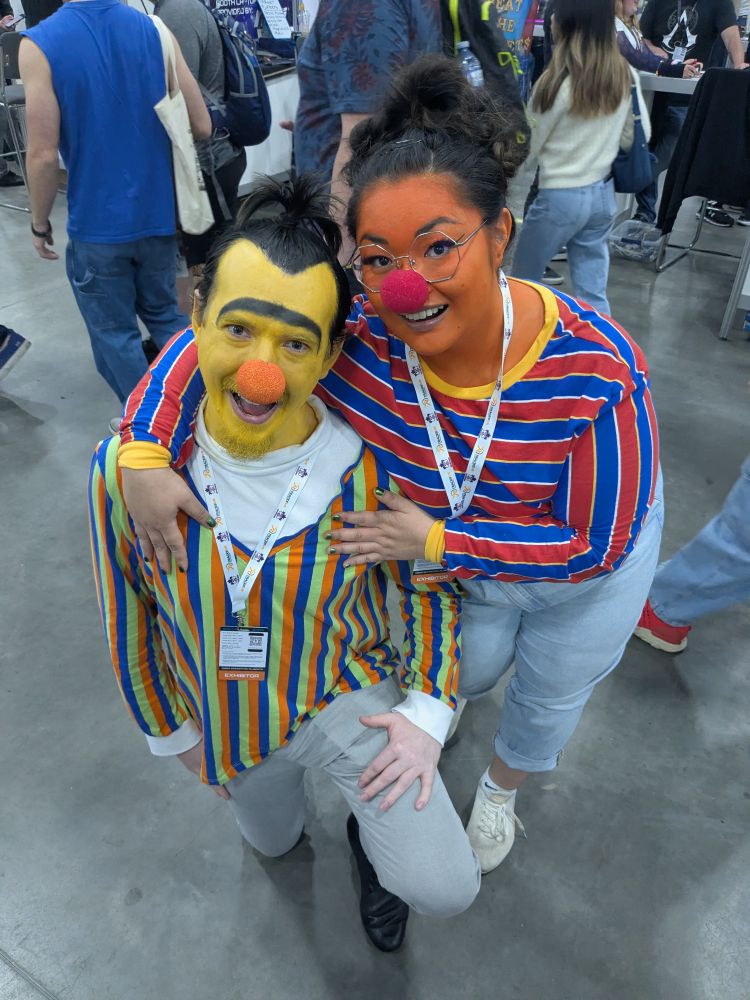A man and a woman cosplaying as Bert and Ernie from Sesame Street. The man dressed as Bert is wearing an orange, green, and blue vertical striped long sleeve shirt over a white turtleneck with grey pants. The woman dressed as Ernie is wearing a red, blue, and white horizontally striped long sleeved shirt with a yellow collar. The man is kneeling on one knee facing the camera, and the woman is crouched down with her arms around the man. Both are looking at the camera and smiling. 