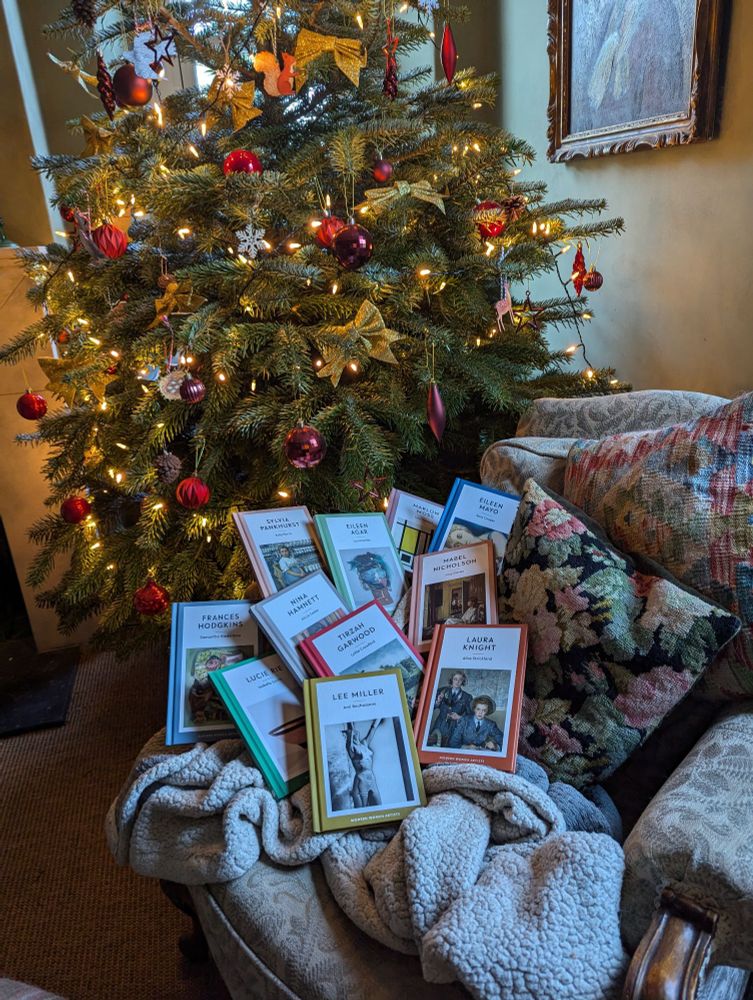 A photograph of a group of colourful art books displayed on a green and cream armchair with tapestry cushions in front of a Christmas tree decorated with with fairy lights, good bows, red baubles in a green living room 