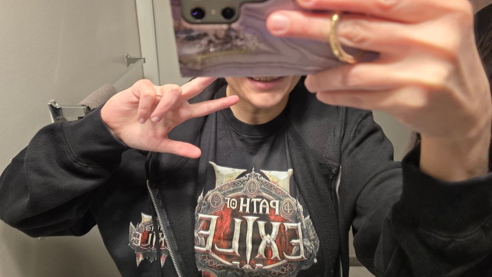 Bathroom mirror selfie of a white female wearing a Path of Exile 2 zip-up hoodie over a Path of Exile 2 t-shirt. Her phone is obscuring most of her face, with just a small bit of the lower portion visible, showing half of a smile. She is making a peace sign with her free hand. 