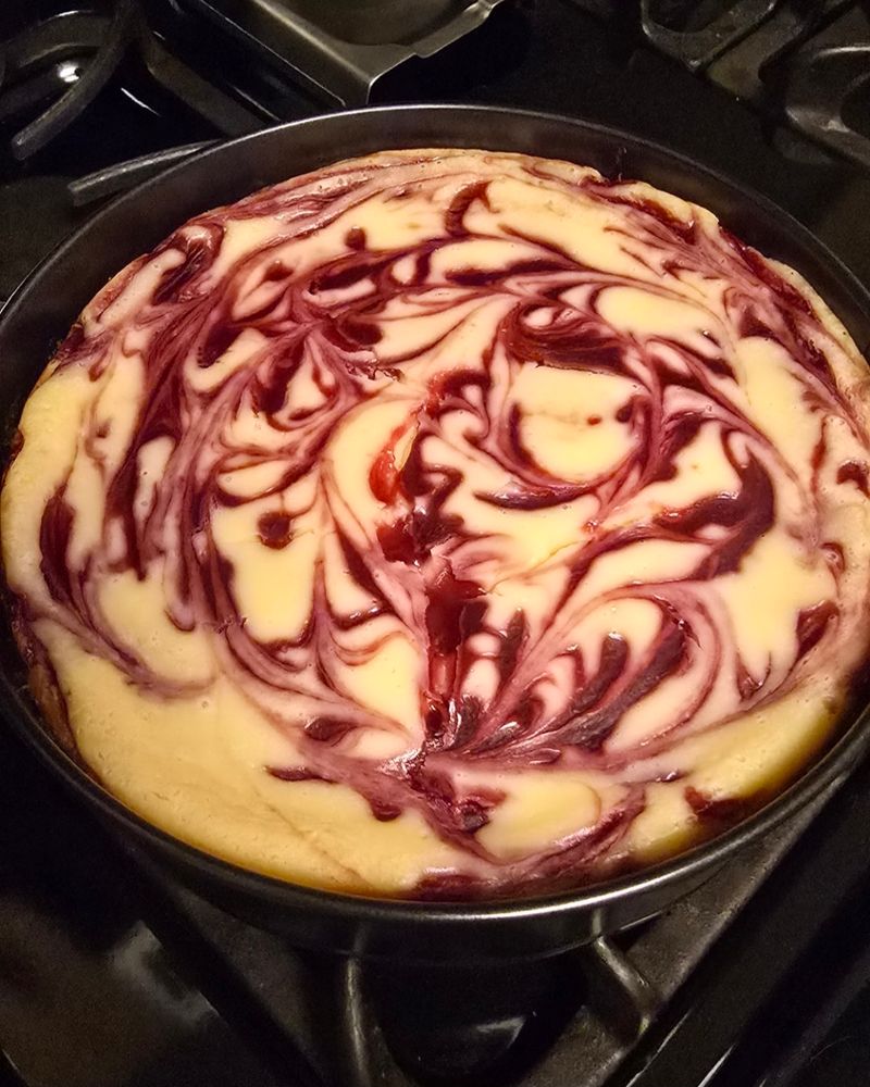 A photo of a cheesecake. There are red swirls throughout the top of it, and it is showing some small signs of cracking. The baker has not been able to prevent this in 9+ years of making this recipe.