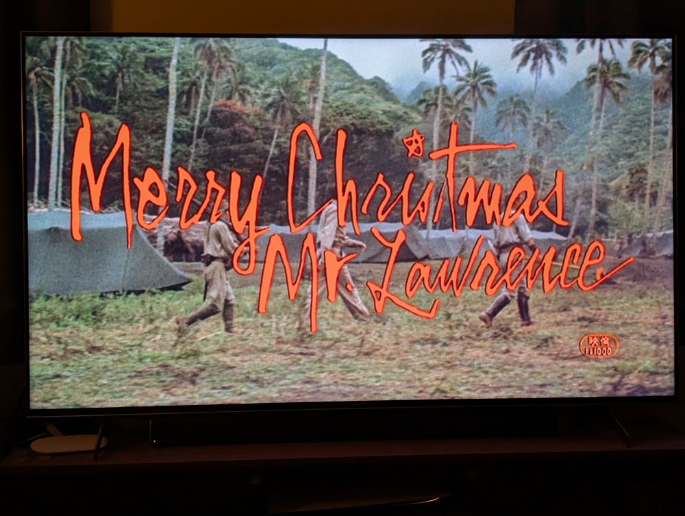 Title screen for Merry Christmas Mr Lawrence. Orange text over a jungle camp 