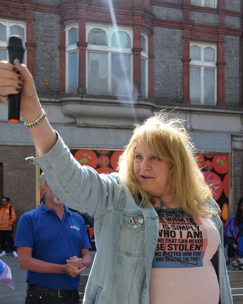 A woman with long blonde hair wearing a denim jacket and a t-shirt with pro-trans rights slogans on it. She is standing in front of a public protest, defiantly holding up a microphone.