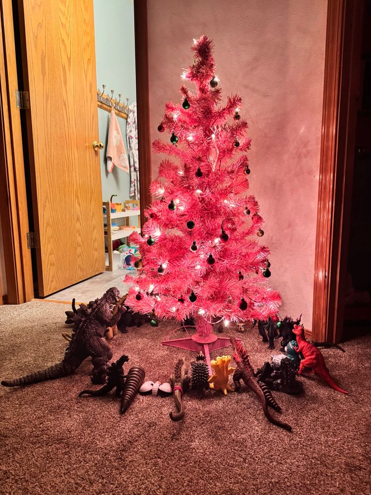 A small, pink, artificial tree with white lights and small red and green ornaments. Encircling the tree are 27 Godzilla toys, including multiple versions of Godzilla, Anguirus, Ghidorah, and Mothra (larval and adult forms). There is also a plastic t-rex named Roar who is not a Godzilla, but who has been invited to live on Monster Island. Roar only says “Roar.”