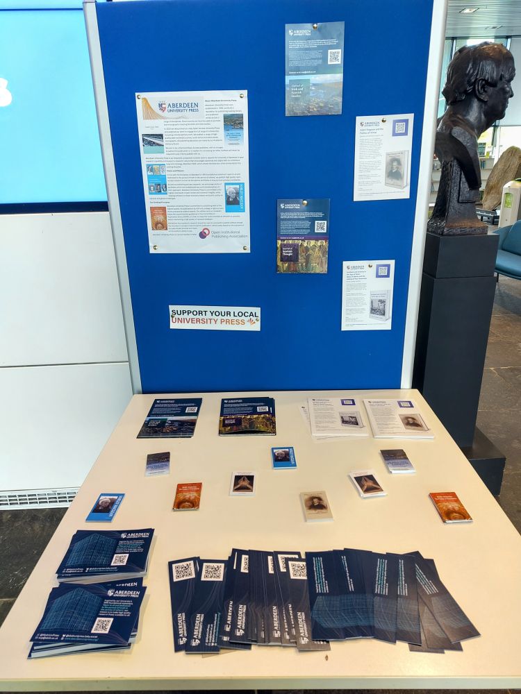 A posterboard with posters and flyers advertising AUP publications, with a table in front full of bookmarks, flyers, and book cards for people to take