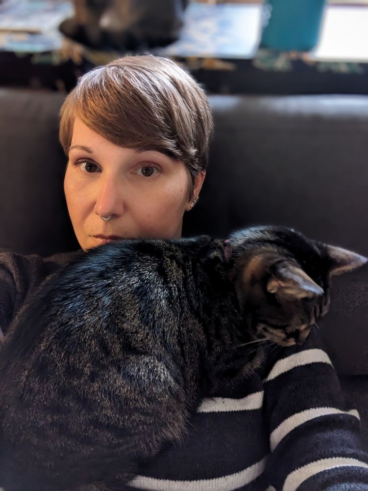 White woman with a pixie cut solemnly looks at the camera while a tabby cat lays on her chest.
